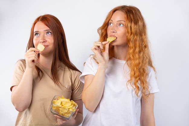 Les filles aux cheveux roux mangent des chips et regardent ailleurs. Photo sur mur blanc. concept de morsure rapide.