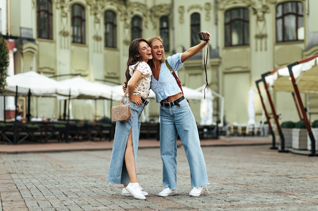 Des filles bronzées joyeuses et heureuses s'embrassent et prennent un selfie à l'extérieur avec une caméra rétro