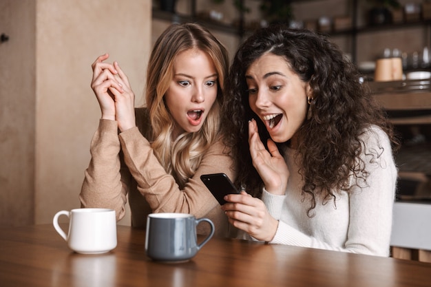 Filles choquées émotionnelles excitées amies assises dans un café à l'aide d'un téléphone portable