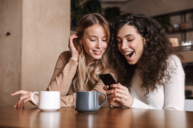 Filles choquées émotionnelles excitées amies assises dans un café à l'aide d'un téléphone portable