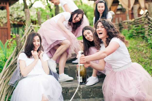 Des filles gaies et une jeune mariée en t-shirts blancs et jupes en tulle rose et moelleux, crachées au moment de l'ouverture du champagne, de la mousse sort de la bouteille, des filles rient et se cachent. Célébration de la fête de poule