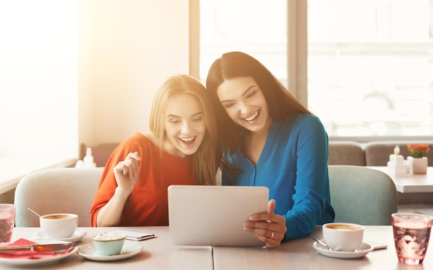 Filles heureuses avec tablette au restaurant. Amis discutant en ligne sur un appareil numérique au café