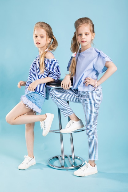 Des filles jumelles dans des vêtements bleu clair posent près d'un tabouret de bar sur un bleu.