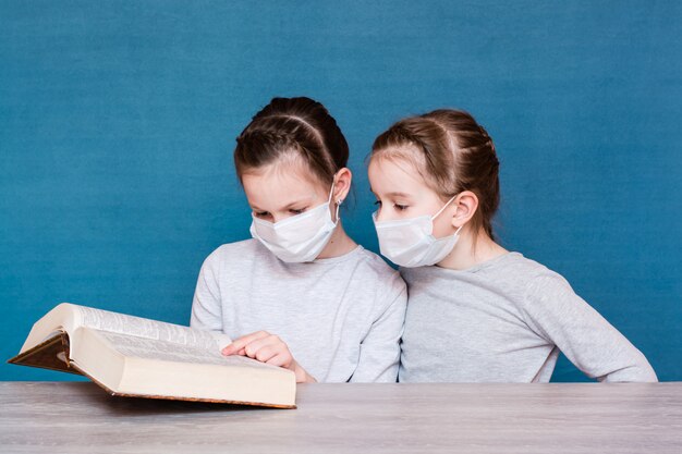 Des filles en masque médical en quarantaine lisent un livre à table. Éduquer les enfants isolés dans une épidémie