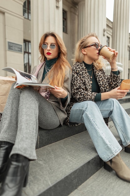 Filles à la mode dans des vêtements à la mode lisant un magazine et buvant du café dans la ville