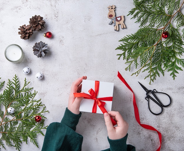 Filles ouvrant le cadeau de Noël blanc avec ruban rouge sur table gris clair. Fond de Noël avec arbre de thuya, cônes de bougies et jouets de Noël