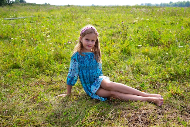 Une fillette de 10 ans vêtue d'une robe bleue est assise sur un champ d'herbe et de fleurs, longues jambes nues, pieds nus