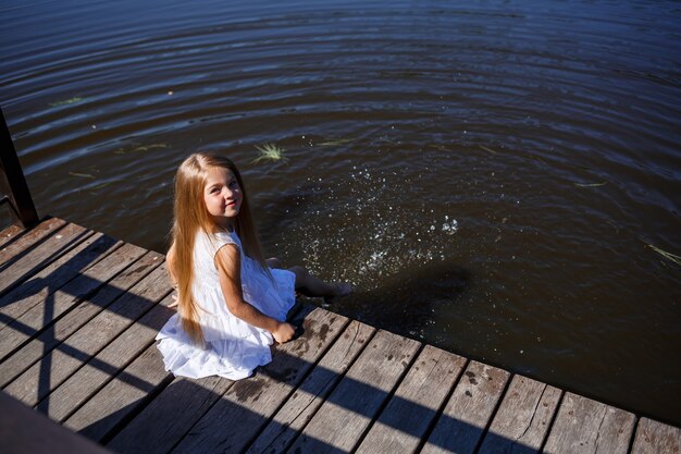 Une fillette de 7 ans aux longs cheveux blonds au bord du lac est assise sur une pochette avec les jambes dans l'eau. Elle éclabousse ses pieds dans le lac. Fille aux pieds nus dans une robe blanche aux cheveux longs.