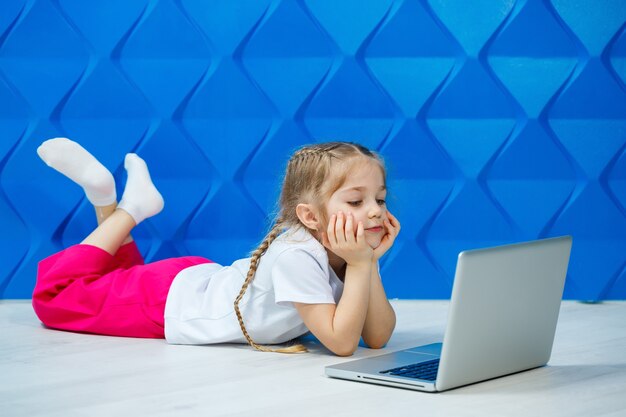 Une fillette de 7 ans en T-shirt blanc est assise par terre avec un ordinateur portable et appuie sur les touches