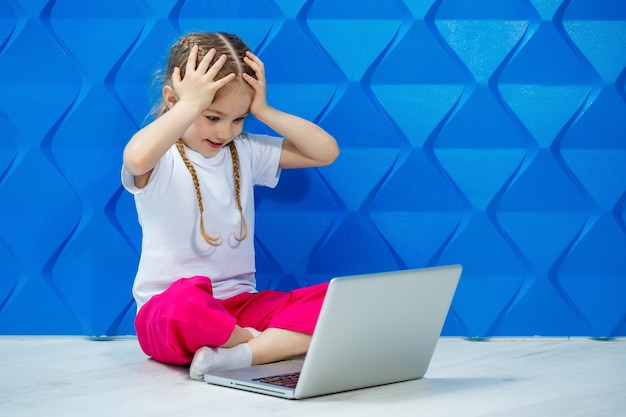 Une fillette de 7 ans en T-shirt blanc est assise par terre avec un ordinateur portable et appuie sur les touches