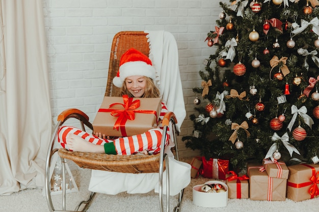 Une fillette de 9 ans tenant un cadeau du Père Noël et assise sur une chaise berçante avec des guirlandes en arrière-plan. Période de Noël. Bonne année!