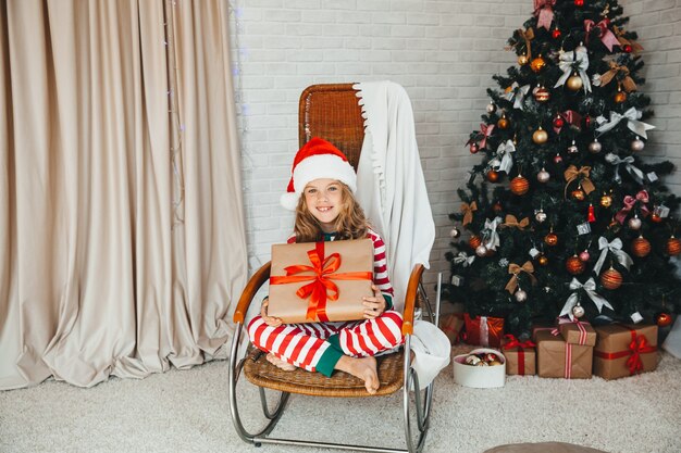 Une fillette de 9 ans tenant un cadeau du Père Noël et assise sur une chaise berçante avec des guirlandes en arrière-plan. Période de Noël. Bonne année!