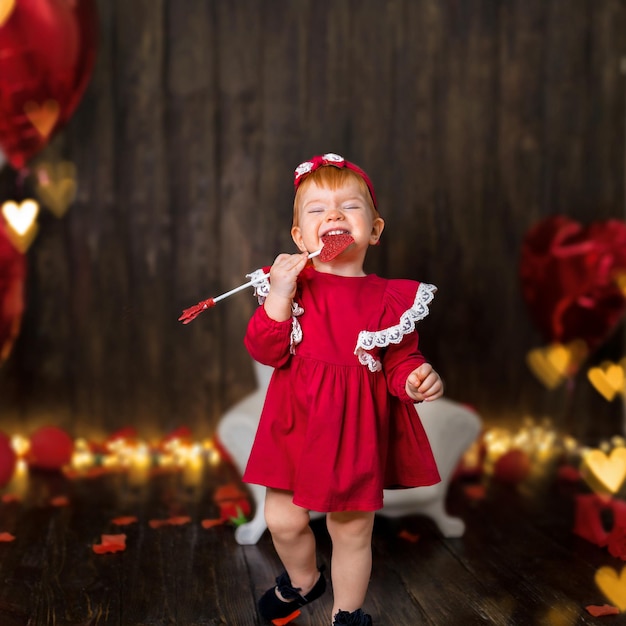 Une fillette d'un an avec une émotion heureuse dans une robe rouge avec une flèche de Cupidon sur un fond en bois sombre...