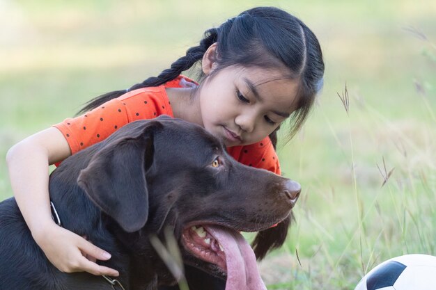 Une fillette d'Asie du Sud-Est avec le football avec son gros chien noir à l'extérieur du terrain en herbe dans l'arrière-cour le soir. Concept d'amoureux des animaux de compagnie