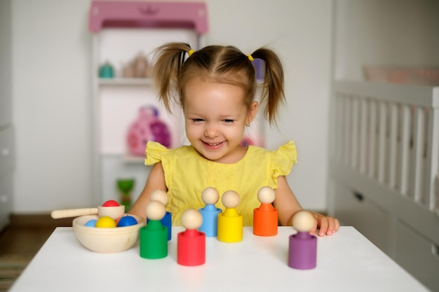 Une fillette de deux ans avec des queues de cheval est assise à une table avec des jouets de développement et rit