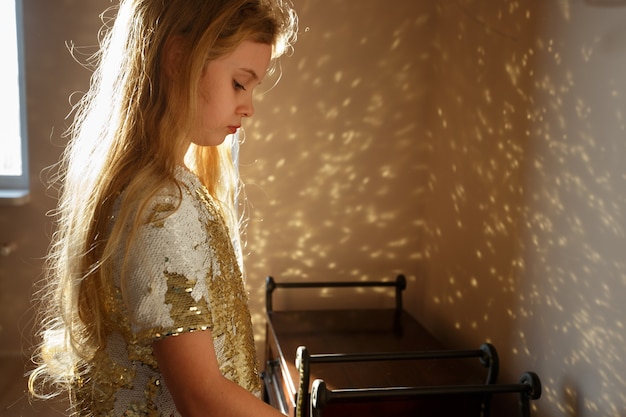 Une fillette de sept ans vêtue d'une robe élégante décorée de paillettes dorées se tient dans la pièce, la lumière du soleil se reflète sur la robe et jette un éclat sur le mur.