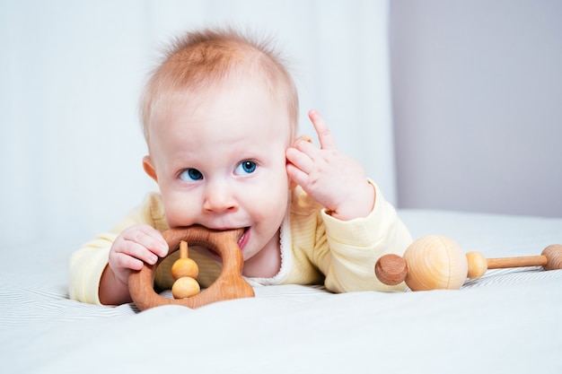 Une fillette de sept mois aux yeux bleus est allongée sur le ventre dans une pièce lumineuse et grignote un jouet en bois en levant les yeux. Les dents de l'enfant font ses dents, les gencives démangent. Concept de la santé des enfants.