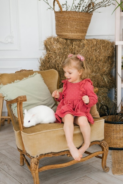 Une fillette de trois ans vêtue d'une robe pieds nus est assise sur une chaise avec un lapin blanc dans le salon décoré pour Pâques