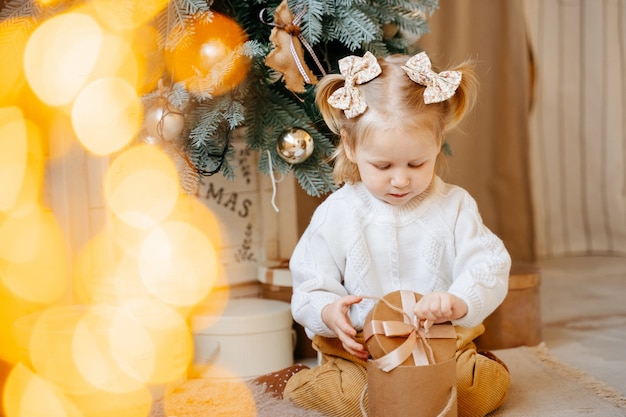 Une fillette vêtue d'un pull blanc est assise près de l'arbre de Noël dans le salon et ouvre une boîte avec un cadeau. Concept de vacances en famille.