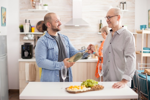 Fils souriant à son père dans la cuisine tenant une bouteille de vin tandis que la mère et la femme sont occupées avec un délicieux brunch. préparation.