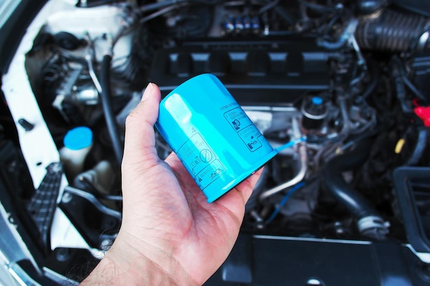 Le filtre à huile moteur automobile tient dans la main du mécanicien automobile avec un compartiment moteur de voiture sur le concept de service de maintenance automatique en arrière-plan