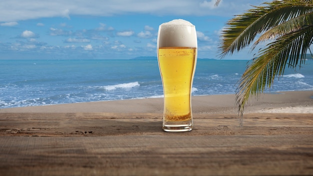 Fin de semaine. Verre avec de la bière froide au soleil avec la mer ou la plage de l'océan avec des palmiers et du sable blanc en arrière-plan. Boisson alcoolisée traditionnelle sur table en bois. Concept d'été, de vacances, de villégiature, de détente.