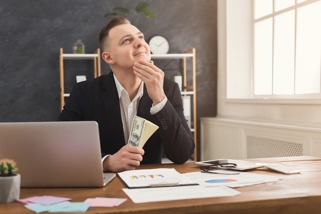 Un financier masculin réfléchi assis à une table de bureau, tenant de l'argent dans les mains et rêvant. Homme comptant son revenu, copiez l'espace. Le profit et les rêves deviennent réalité concept