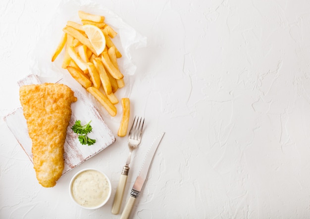 Photo fish and chips britannique traditionnel avec sauce tartare sur une planche à découper avec une fourchette et un couteau et une tranche de citron sur une table en pierre blanche.