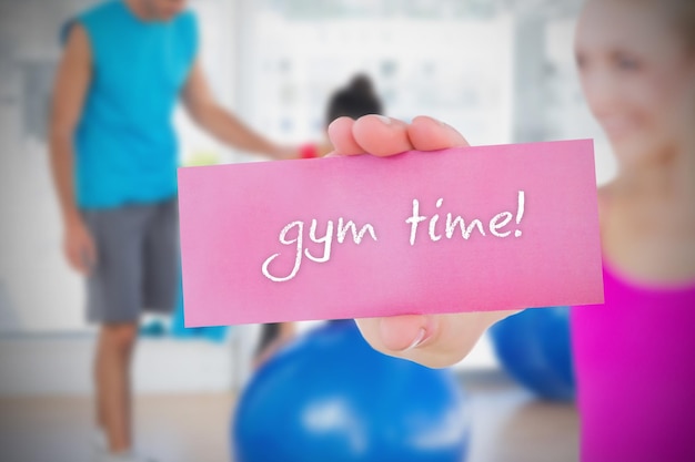 Fit blonde holding card disant temps de gym contre l'entraîneur et le client dans un studio de fitness