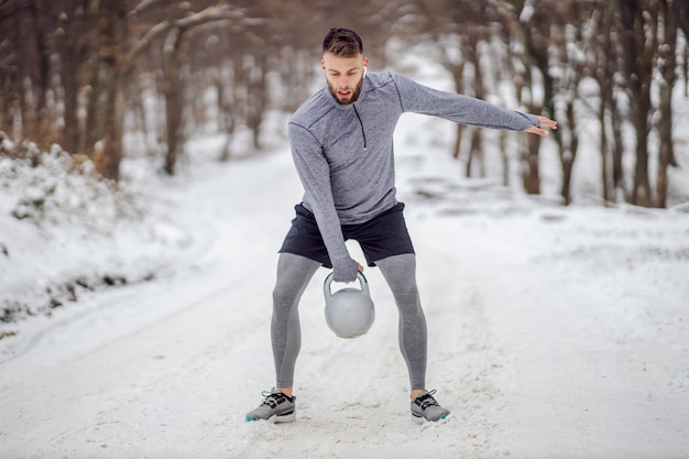 Fit bodybuilder debout sur un chemin enneigé en forêt et balançant kettlebell en hiver. Exercices de musculation, fitness hiver, fitness en plein air