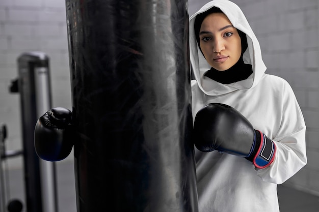 Fit femme arabe prenant une pause après une formation de boxe dure, une femme musulmane se tient près de sac de boxe posant à la caméra, dans une salle de sport. femme en hijab