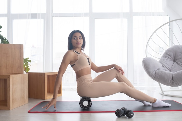 Fit femme noire sportive portant des vêtements de sport rouleau sur rouleau de massage myofascial mousse massage musculaire sur tapis de yoga à la maison