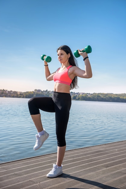 Photo fit girl faisant de l'exercice avec des haltères dans ses mains à la nature, près du lac