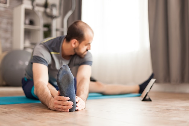 Fit un homme étirant son corps en regardant un cours de pilates en ligne sur une tablette pendant le verrouillage mondial.