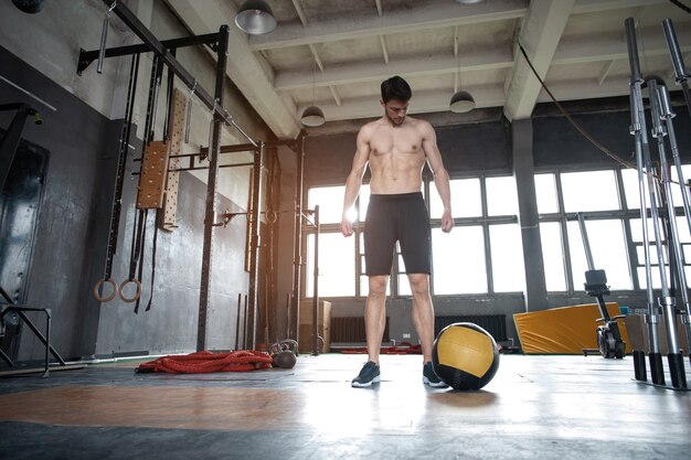 Fit l'homme lançant un ballon de médecine faisant un coup de balle contre le sol de la salle de sport ou un exercice d'entraînement du haut du corps avec une pression sur les épaules. Entraînement croisé au centre de remise en forme.