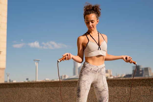 Fit jeune femme active avec corde à sauter l'exercice à l'extérieur le matin contre le ciel bleu et l'architecture moderne