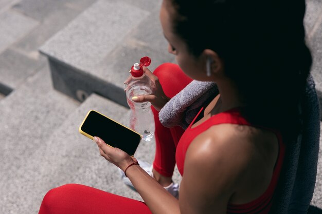 Fit jeune femme aux cheveux noirs en vêtements de sport rouges et écouteurs blancs détient smartphone et bouteille d'eau. Vue de coupe à angle élevé. Une serviette violette est accrochée à son épaule. Sport dans le concept de grande ville.