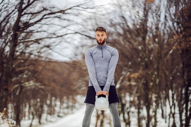 Fit sportif debout sur un chemin enneigé en forêt et balançant kettlebell en hiver. Exercices de musculation, fitness hiver, fitness en plein air