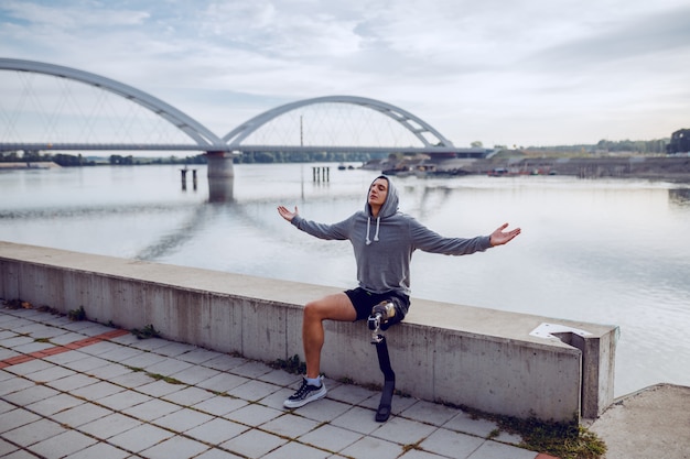 Fit sportif handicapé caucasien sain avec jambe artificielle et sweat à capuche sur la tête assis sur le quai avec les bras ouverts.