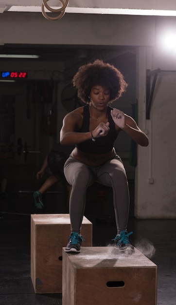 Photo fit young african american woman box jumping dans une salle de sport de style crossfit. l'athlète féminine effectue des sauts de boîte au gymnase.