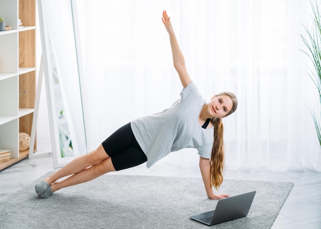 Fitness à domicile pour enfants Pilates en ligne Entraînement matinal Mode de vie actif Fille athlétique en t-shirt tenant une planche latérale seule avec un didacticiel de sport vidéo sur ordinateur portable à l'intérieur de la pièce lumineuse