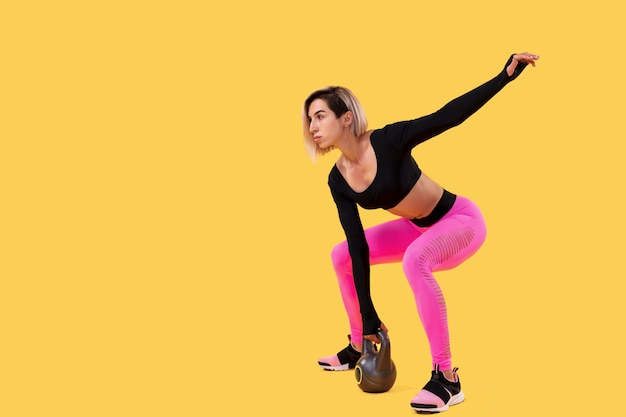 Fitness femme en sportswear rose et noir à la mode travailler avec kettlebell sur mur jaune. Force et motivation.