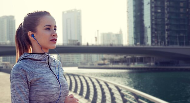 fitness, sport, gens, technologie et style de vie concept - femme heureuse courant et écoutant de la musique dans des écouteurs sur le fond de la rue de la ville de Dubaï