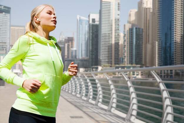 fitness, sport, personnes et concept de mode de vie sain - jeune femme avec des écouteurs faisant du jogging sur la rue de la ville de dubaï ou sur le front de mer