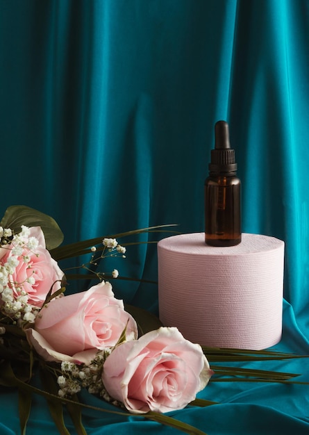 Flacon compte-gouttes en verre sur un podium avec des fleurs roses roses Fond de rideau de soie bleu Concept de beauté