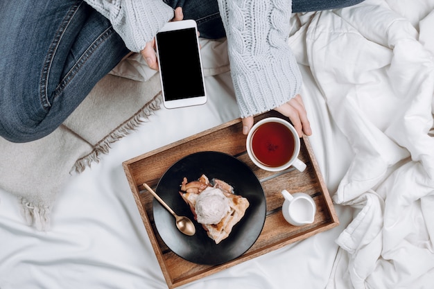 Flatlay confortable de lit avec plateau en bois avec tarte aux pommes végétalienne, crème glacée et thé noir et femme en jeans et pull gris tenant un smartphone avec fond noir sur des draps blancs et des couvertures