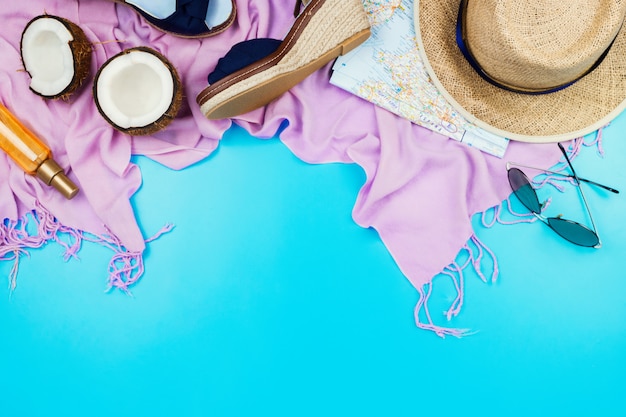 Flatlay de vacances d'été avec chapeau de paille, écharpe rose, espadrilles, noix de coco, huile pour le corps et verres sur fond bleu avec fond