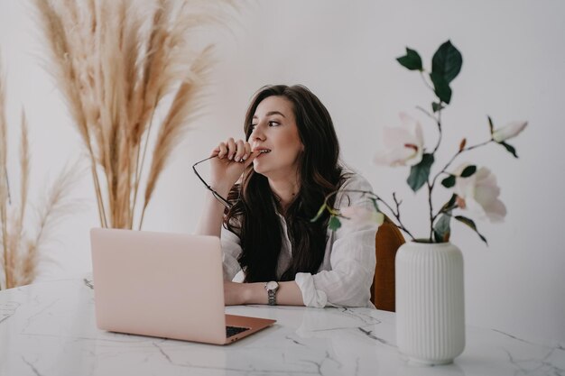 Fleuriste designer belle femme brune assise au bureau regardant de côté avec des lunettes ii sa main devant un ordinateur portable à table vêtue d'une chemise blanche contre des plantes floues Concept de personnes créatives