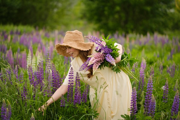 Un fleuriste heureux vêtu d'une robe jaune et d'un chapeau de paille cueille des fleurs de lupin violet dans un champ