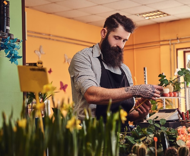 Fleuriste masculin professionnel avec barbe et tatouage sur sa main faisant une belle composition de fleurs tout en se tenant au comptoir d'un magasin de fleurs.